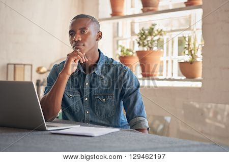 Handsome young designer of African descent sitting in his beautifully lit studio, looking thoughtfully into the distance with his laptop open in front of him