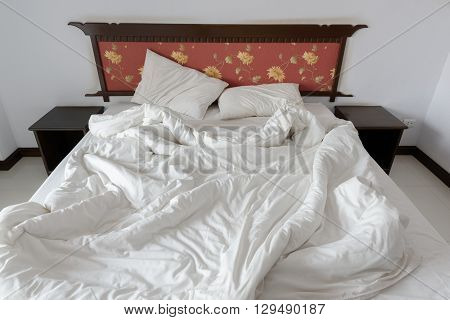 Untidy / unmade bed with two messy pillows and a white crumpled blanket in a hotel single (standard) room. Bedclothes are not neatly arranged for a new customer / guest to check in and sleep in.