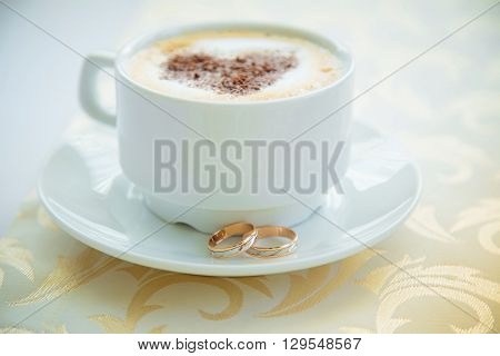 Wedding day. White cup of cappuccino with a painted heart of chocolate and a pair of gold wedding rings on a white saucer