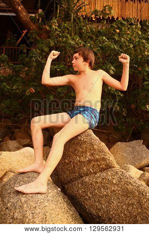 preteen hadsome suntanned boy in swimming slop show biceps on the stone rock beack background close up photo