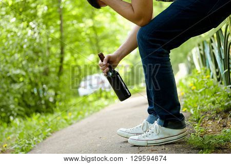 Man depressed with beer bottle sitting on green fence outdoor. 