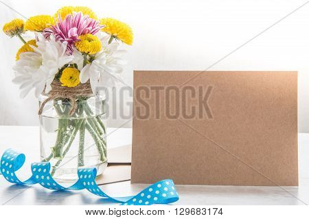 Spring flowers bouquet in a jar vase with kraft card note envelope and blue dotted ribbon on a white wood rustic background