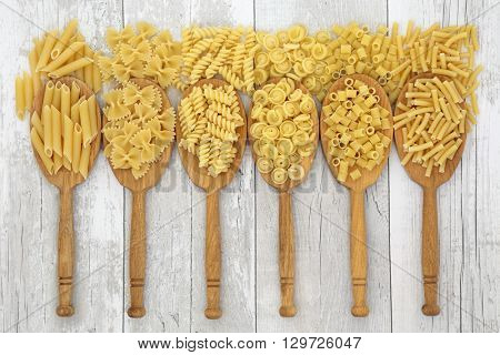 Dried pasta italian food in oak wood spoons and loose over distressed white wood background. Penne, farfalle, fusilli, messicani, ditali rigati and macaroni varieties from left to right. 