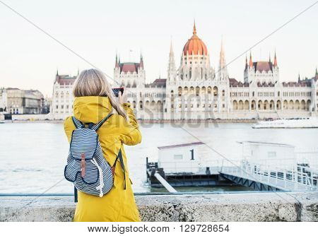 Young blondy woman tourist making photos of Parliament historic building from Danube side with her phone in Budapest, Hungary, on a spring clear day