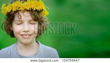 Handsome boy with a wreath of yellow dandelions on his head closed his eyes, breathing warm air summer.