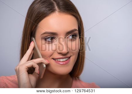 Involved in conversation.  Portrait of  cheerful delighted woman smiling and talking on cell phone while standing isolated on grey background