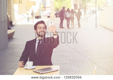 Toned image of handsome businessman making selfies on his mobile or smart phone while resting or working in restaurant or cafe in city centre.