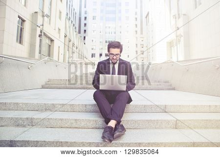 Toned image of handsome businessman sitting on stairs incity centre and using laptop computer. Freelance man in glasses and black business suit working.
