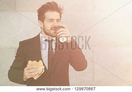 Closeuptoned picture of businessman eating junk food and drinkig tea or coffee while walking to office along street. Happy freelancer in hurry.