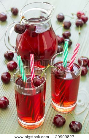 Two glasses of cherry juice with straws in the foreground and in the background a jar full of cherry juice. 2 cherry juices. Vertical. Close-up. Daylight.
