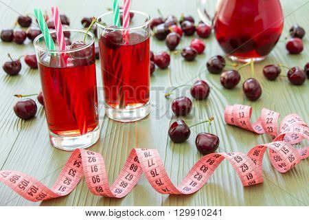 Left 2 glasses of juice with straws, scattered cherries, jar of cherry juice, in front centimeter on light green background. Cherries, cherry juice and centimeter. Horizontal.