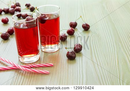 Left two glasses of cherry juice around scattered cherries, right empty space for text on light green background.  2 cherry juices, straws and empty space. Horizontal. Close. Daylight.