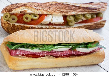 Horizontally in the foreground lying sandwich with salami, mozzarella, arugula, tomatoes and behind sandwich with feta cheese, olives, tomatoes on the light wood background. Horizontal. Daylight. Close.