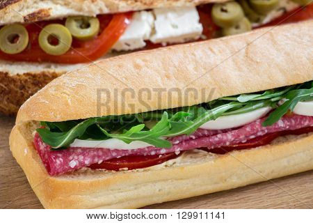 Sandwich with salami, mozzarella, arugula and tomato, and sandwich with feta cheese, olives, tomato on the light wood background. Horizontally one behind the other 2 different sandwiches. Horizontal. Daylight.