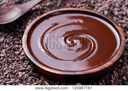 Bowl of melted chocolate and wooden spoon on a crushed raw cocoa beans, nibs background. Copy space.