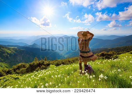 On The Lawn In Mountains Landscapes The Hipster Girl In Dress, Stockings And Straw Hat Stays Watchin