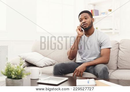 Upset Young African-american Man Talking On Smartphone Emotionaly, Sitting On Beige Couch At Home , 