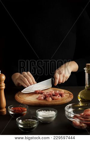 Hands Cut The Raw Meat Into Pieces On Wooden Board On Dark Table With Spices And Bottle Of Oil. Proc