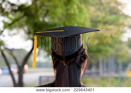 Behind Photo Of University Graduate Wears Gown And Black Cap, Yellow Ribbon And Blur Bokhe Backgroun