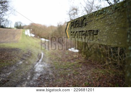 Wooden public footpath sign taken with a shallow depth of field, pointing towards a muddy path with the last signs of snow nestled against a hedgerow on a cold winter morning.