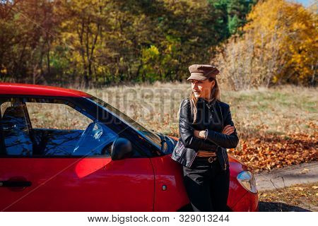 Young Woman Standing By Car On Autumn Road. Driver Stopped Auto In Forest To Enjoy Fall Landscape