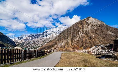 Beautiful Mountain Valley With Stream, Trees, Footpath And Wooden Bridge, Livigno Is A Small Town An