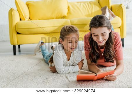 Happy Babysitter Lying On Carpet With Smiling Kid While Reading Book