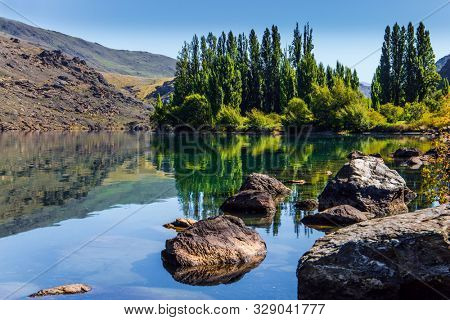 Magic New Zealand. The surroundings of the Cromwell. Ideal place for a picnic. Cypress alley and huge basalt boulders reflected in the lake. The concept of ecological, landscape and photo tourism