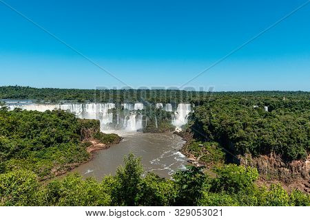 A Green And Lush Rainforest Is Overlooking The Huge Iguazu Falls On A Clear And Sunny Day.