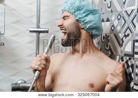Attractive young cheerful man singing while washing in the shower, wearing shower cap and holding shower head