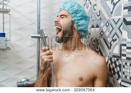 Attractive young cheerful man singing while washing in the shower, wearing shower cap and holding shower head