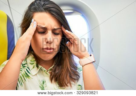 Young traveller woman sitting inside plane at the airport with sky view from the window feeling sick, headache and nauseas