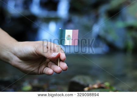 National Flag Of Mexico On Wooden Stick. Young Boy Holds National Athem Near Waterfall On Stream In 
