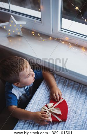 Little Child Boy Holding Red Heart Shaped Gift Box With Bow Near Window In Daylight. Valentine Day P