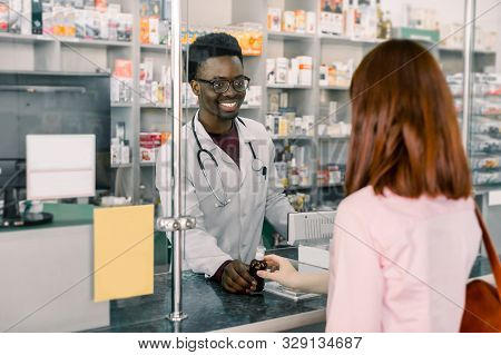 Smiling Male Afrcian Pharmacist Giving Medicine To Young Caucasian Woman In Drugstore