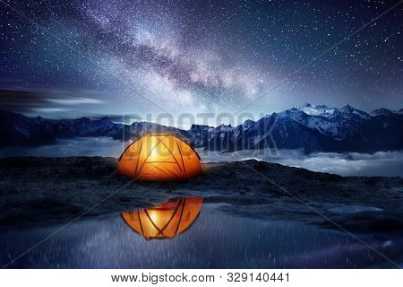Camping Adventure In The Mountains. A Tent Pitched Up And Glowing Under The Milky Way. Photo Composi