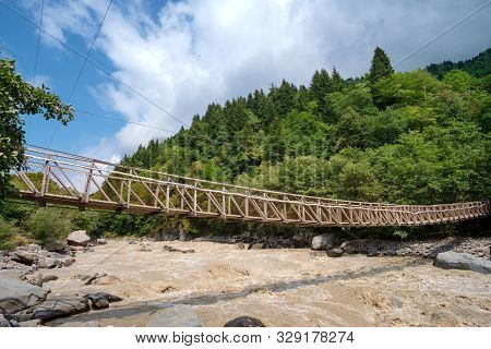Hanged Rope Bridge Over The Fast Muddy River With Green Forest Background