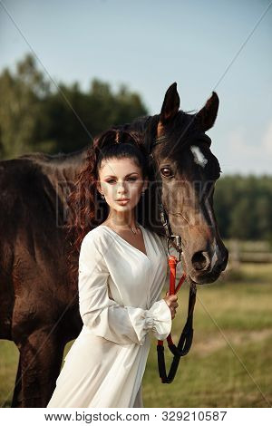 Girl In A Long Dress Stands Near A Horse, A Beautiful Woman Strokes A Horse And Holds The Bridle In
