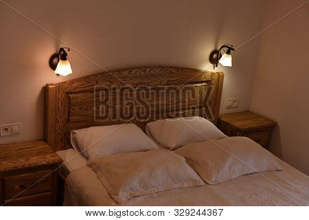 Bed In A Hotel Room. Bed And Pillows. A Bed With A Wooden Headboard And Two Cabinets And Floor Lamps