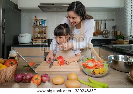 Portrait Of Beautiful Asian Young Woman And Her Daughter Cooking Salad For Lunch Using Online Intern