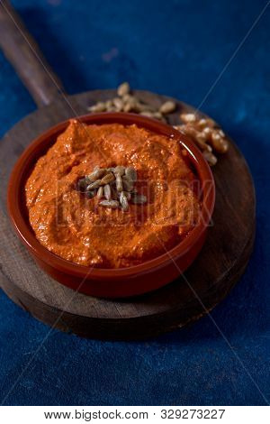 closeup of a brown earthenware bowl with a homemade muhammara, made with red peppers and walnuts, topped with some fried and salted sunflower seeds, on a table