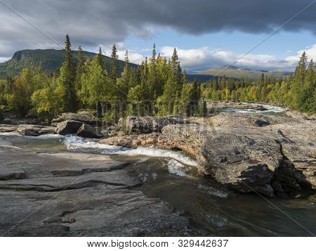 Beautiful Northern Landscape With Wild Glacier River Kamajokk, Boulders And Spruce Tree Forest In Kv