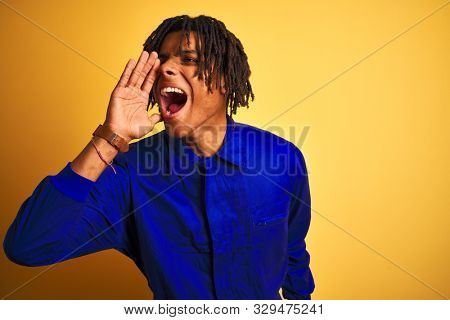Afro worker man with dreadlocks wearing mechanic uniform over isolated yellow background shouting and screaming loud to side with hand on mouth. Communication concept.