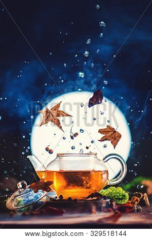 Autumn Tea In Glass Teapot With Shining Full Moon, Falling Leaves And Water Drops. Creative Drink Ph