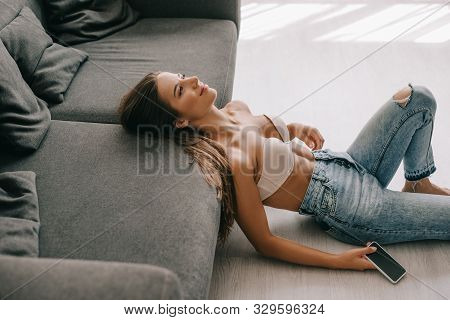 Beautiful Happy Woman In White Bra And Jeans Sitting Near Sofa With Smartphone