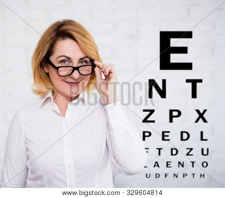 Portrait Of Cheerful Mature Business Woman In Eyeglasses And Eyevision Test Chart Over White Wall Ba