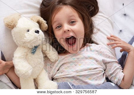 Indoor Shot Of Cute Darkhaired Baby Girl Lying In Bed, Looking Up With Surprised Look And Opened Mou