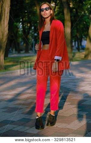 Blonde Girl In A Red Lady-type Pantsuit And Black Top, Watch, Ring, Pendant, Sunglasses Is Walking A