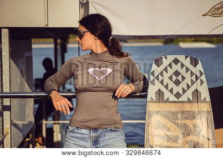 Brunette Girl In Gray Turtleneck, Denim Shorts And Sunglasses Is Posing With Her Wakeboard Satnding 