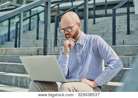 A Bald Guy With A Brown Beard And Pc Glasses Striped Shirt And Beige Pants With Laptop Sitting On Th